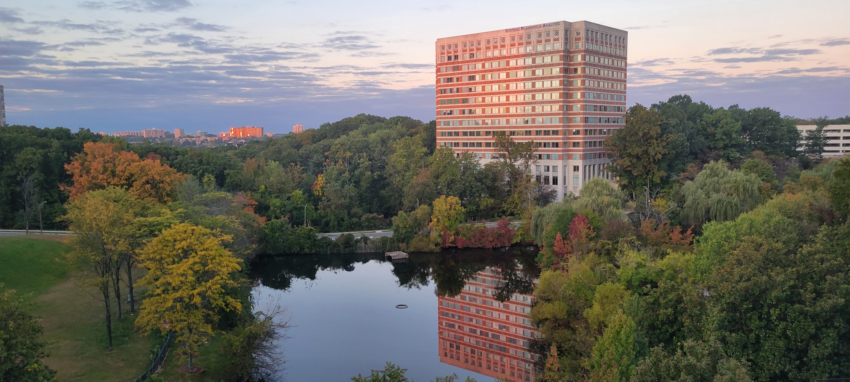 A photo of the Systems Planning and Analysis office building in the background with trees and a scenic lake in the foreground
